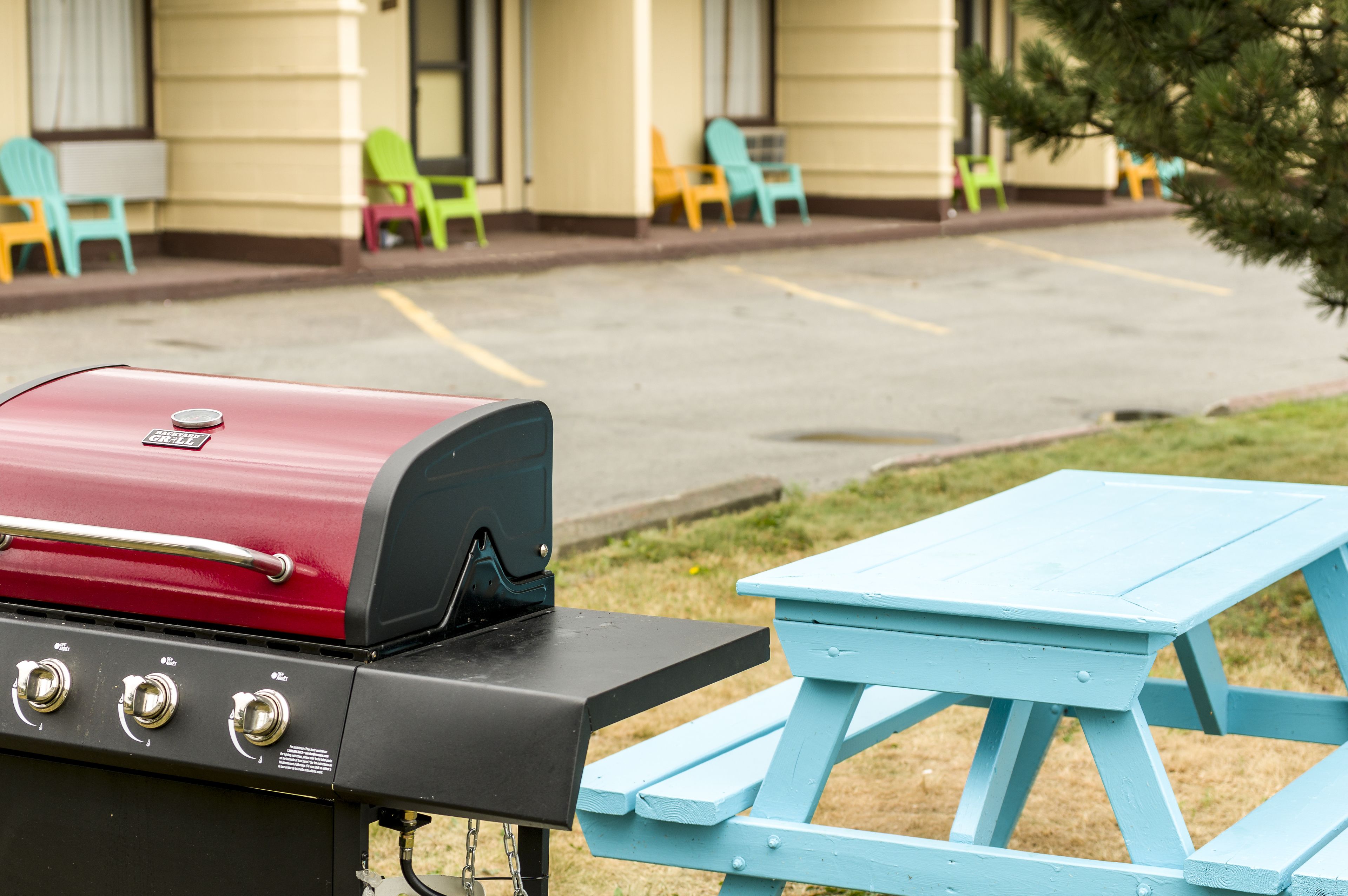 A red barbecue grill with a black lid, a blue picnic table, and colorful chairs in the background.