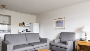 Habitación de hotel moderna con un sofá gris, un sillón a juego, una mesita de madera con lámpara, una obra de arte enmarcada en la pared y una kitchenette al fondo.