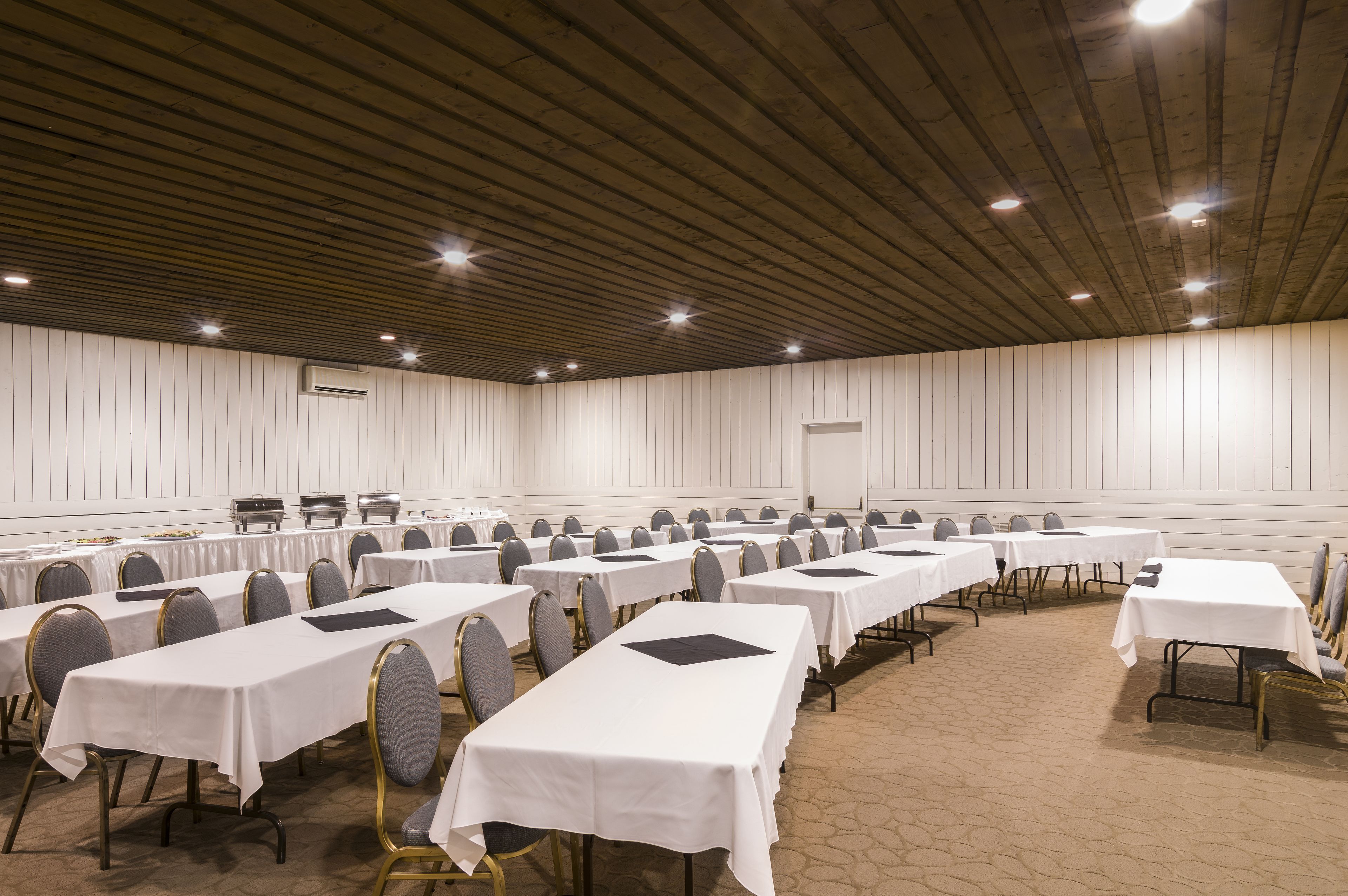 A conference room with multiple tables and chairs, white walls, and a wooden ceiling.