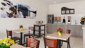 Une cuisine moderne équipée d’une machine à café, d’un réfrigérateur et d’un comptoir où figure une carte des différentes sortes de café. On y trouve deux tables avec des chaises, et un tableau représentant un paysage enneigé est accroché au mur.