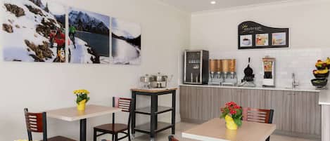 A modern kitchen with a coffee machine, a refrigerator, and a counter with a coffee menu. There are two tables with chairs, and a painting of a snowy landscape on the wall.
