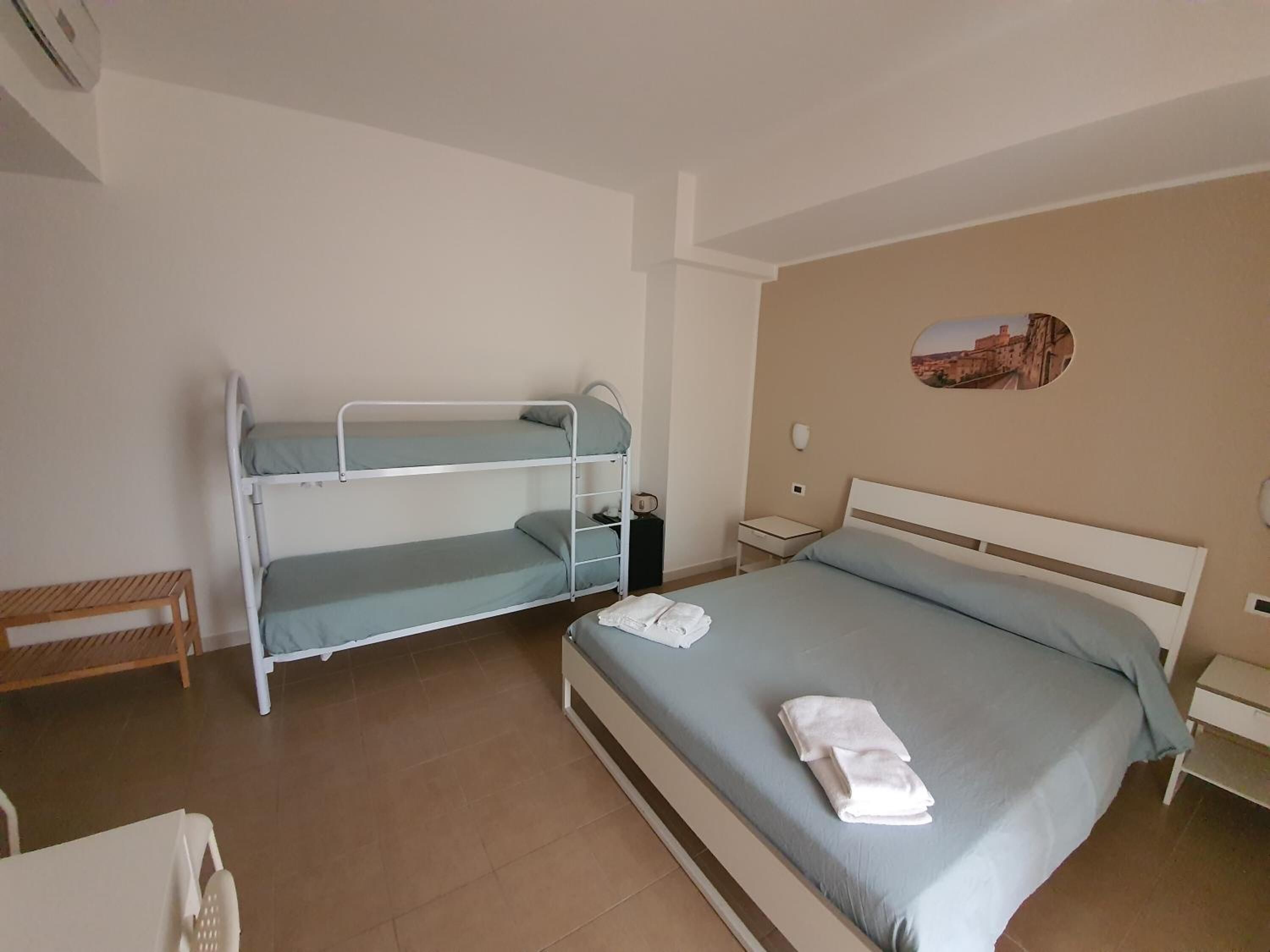 Una habitación con una cama litera, una cama individual, una silla de madera, una mesita de noche y un cuadro colgado en la pared.