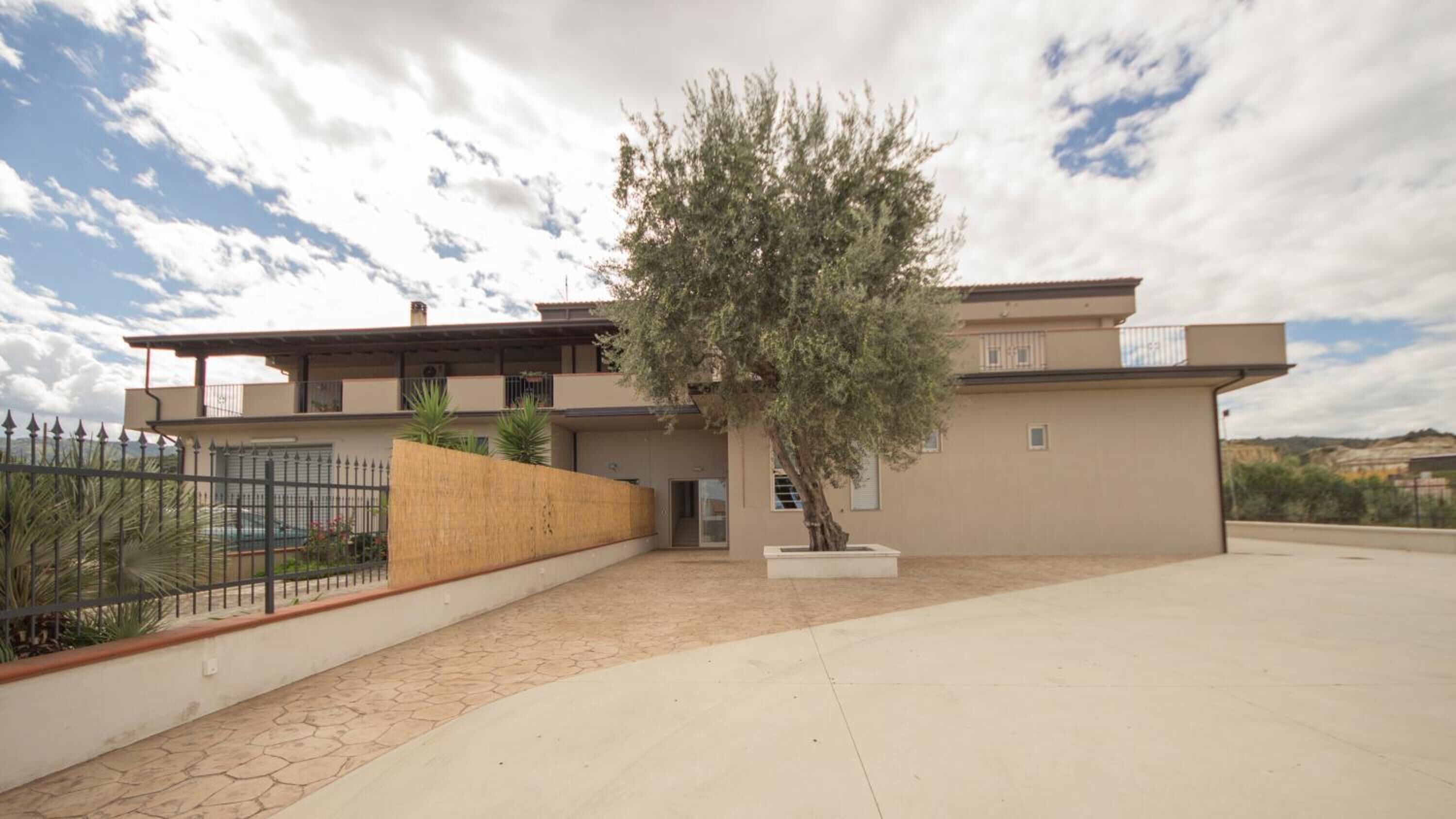 A modern two-story house with a large tree in the front yard, a paved driveway, and a metal fence.