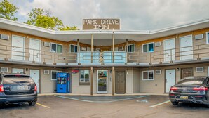 Um motel com uma placa indicando "Park Drive Inn", apresentando uma construção de dois andares com varanda, uma máquina de venda automática de Pepsi e dois carros estacionados.