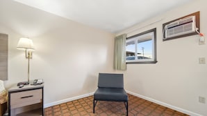 Quarto de hotel com uma cama de solteiro, mesa de cabeceira com abajur, uma cadeira, janela com vista para um edifício e um ar-condicionado fixado na parede.