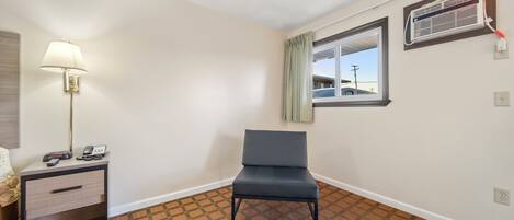 A hotel room with a single bed, a nightstand with a lamp, a chair, a window with a view of a building, and a wall-mounted air conditioning unit.