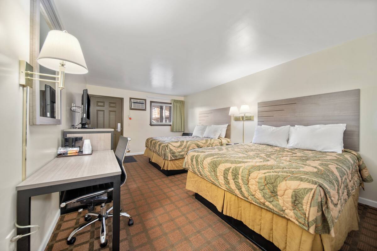 A hotel room with two beds, a desk, and a chair. There is a window with curtains, a lamp, and a mirror.