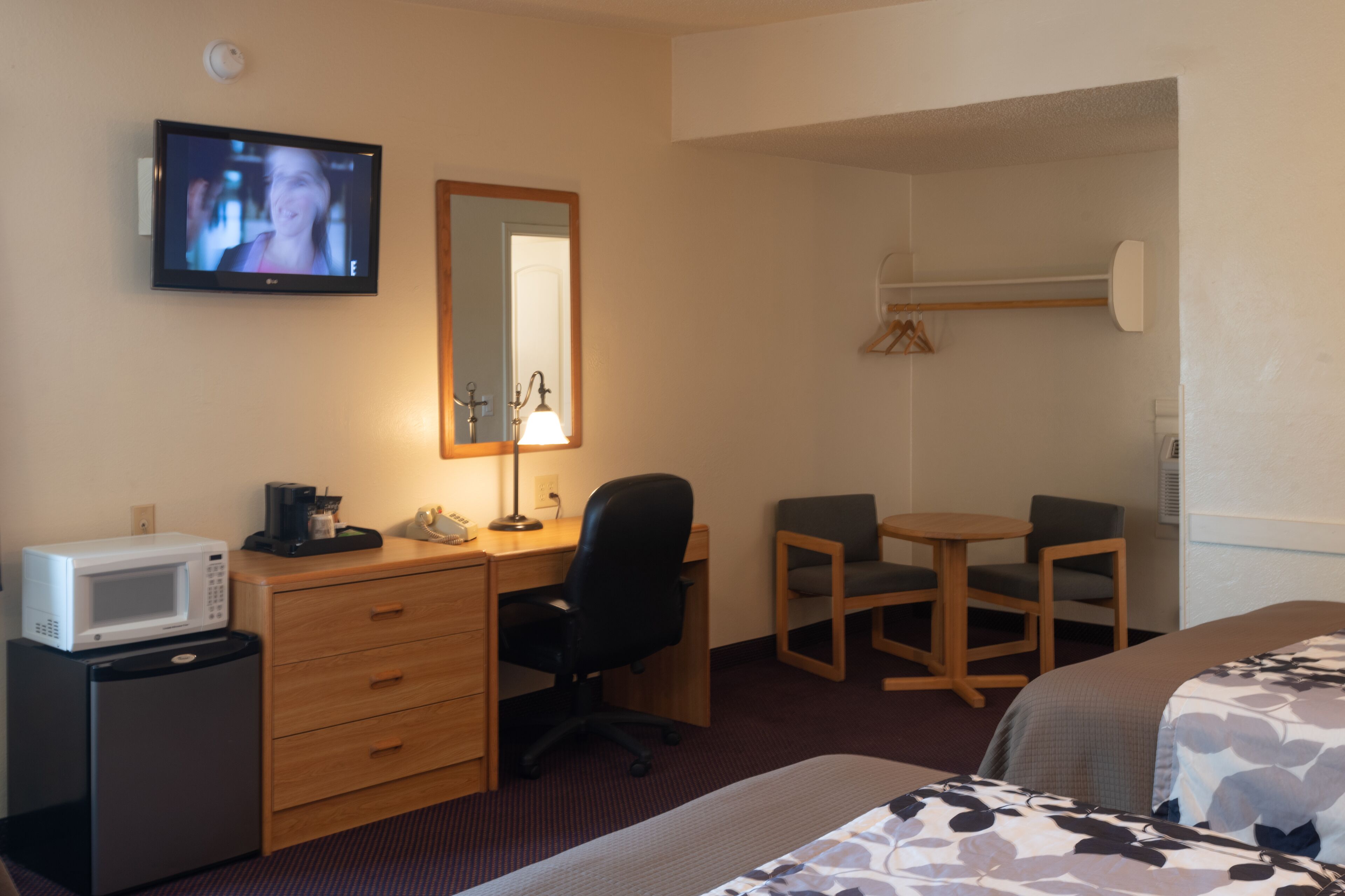 A hotel room with a bed, a desk with a computer, a chair, a small fridge, a microwave, a TV, a lamp, and a mirror.