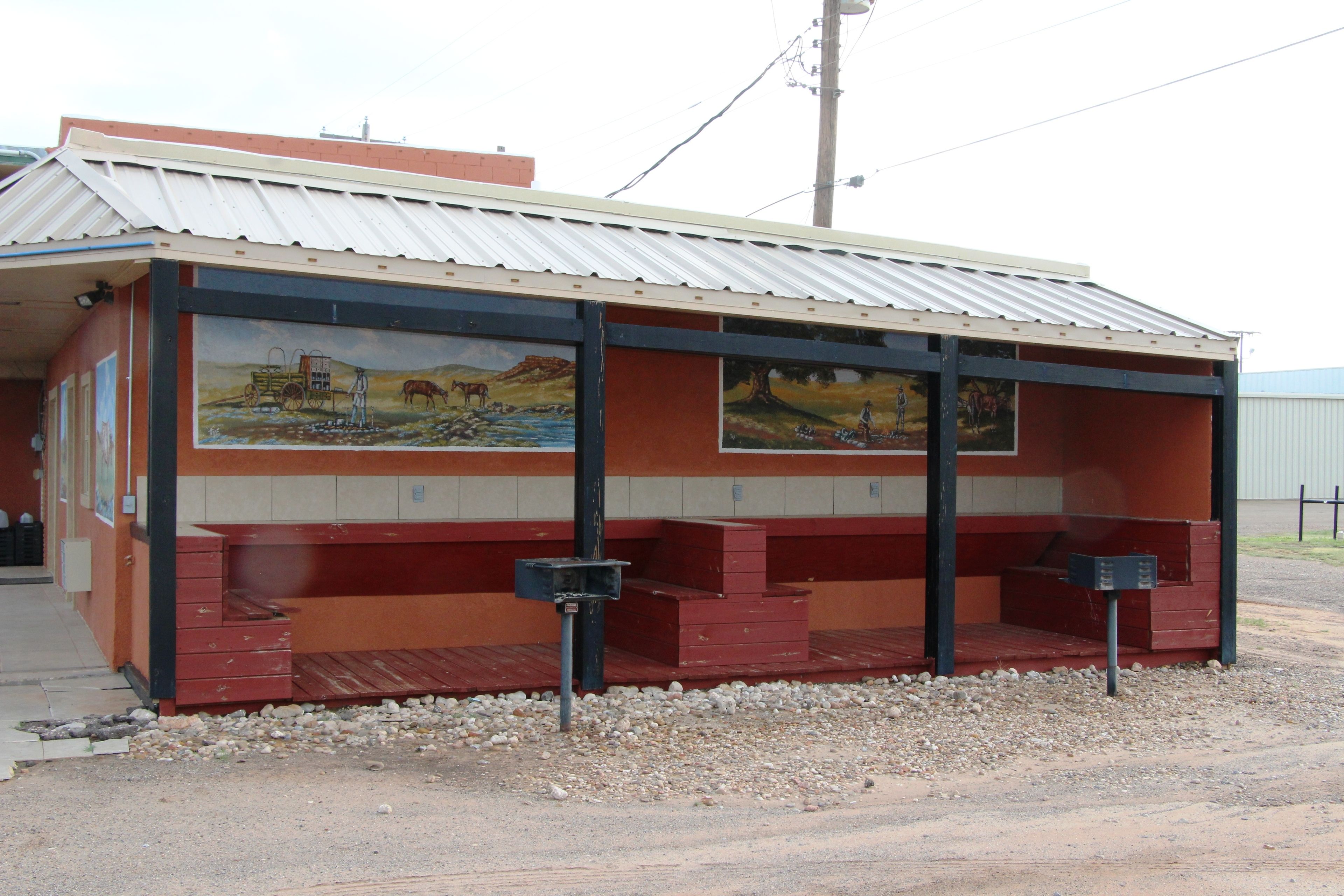 A building with a mural depicting a rural scene, a covered seating area, and a gravel ground.