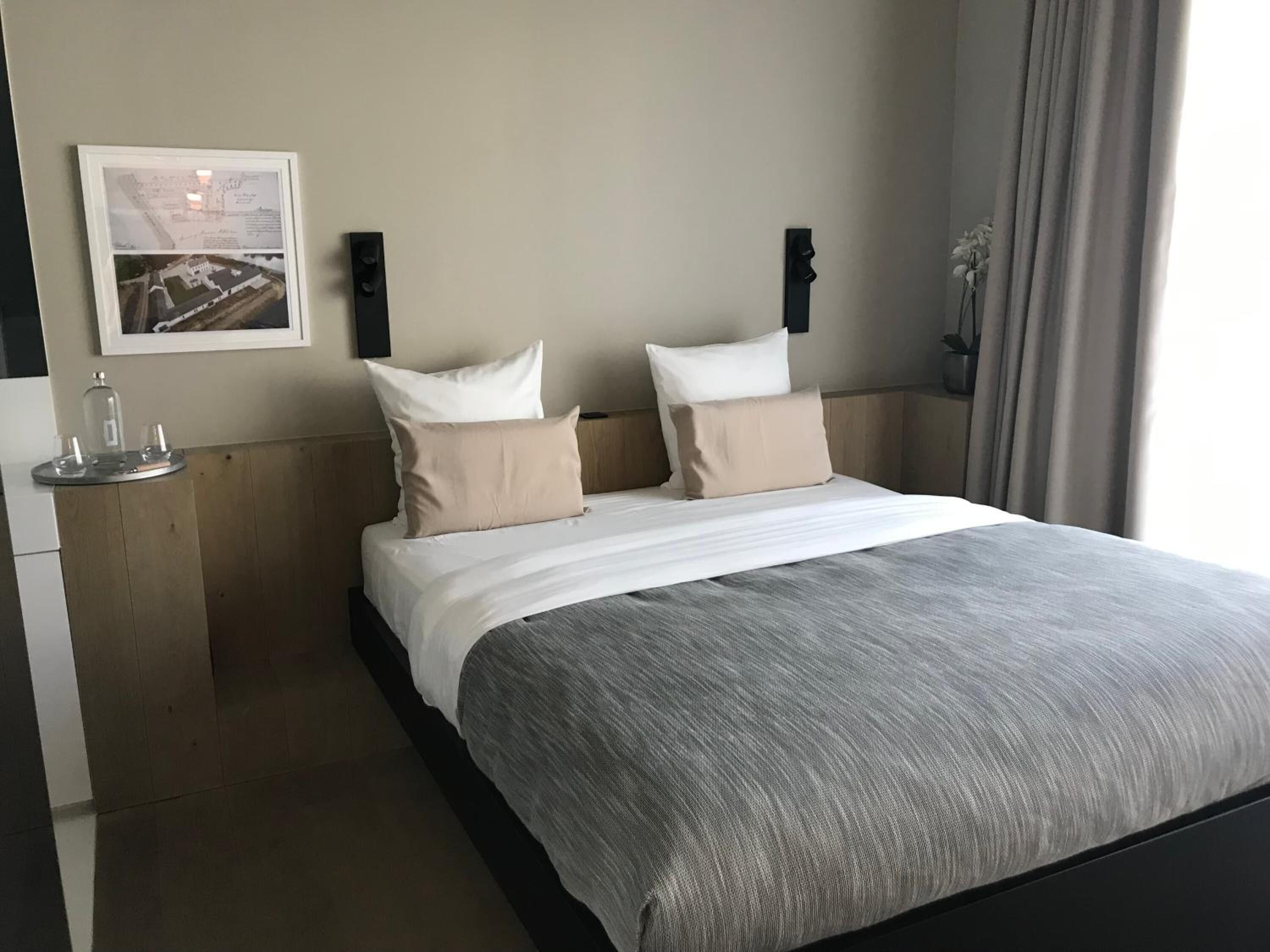 A neatly made bed with a grey blanket and two pillows, a bedside table with a glass decanter and glasses, and a framed map on the wall.