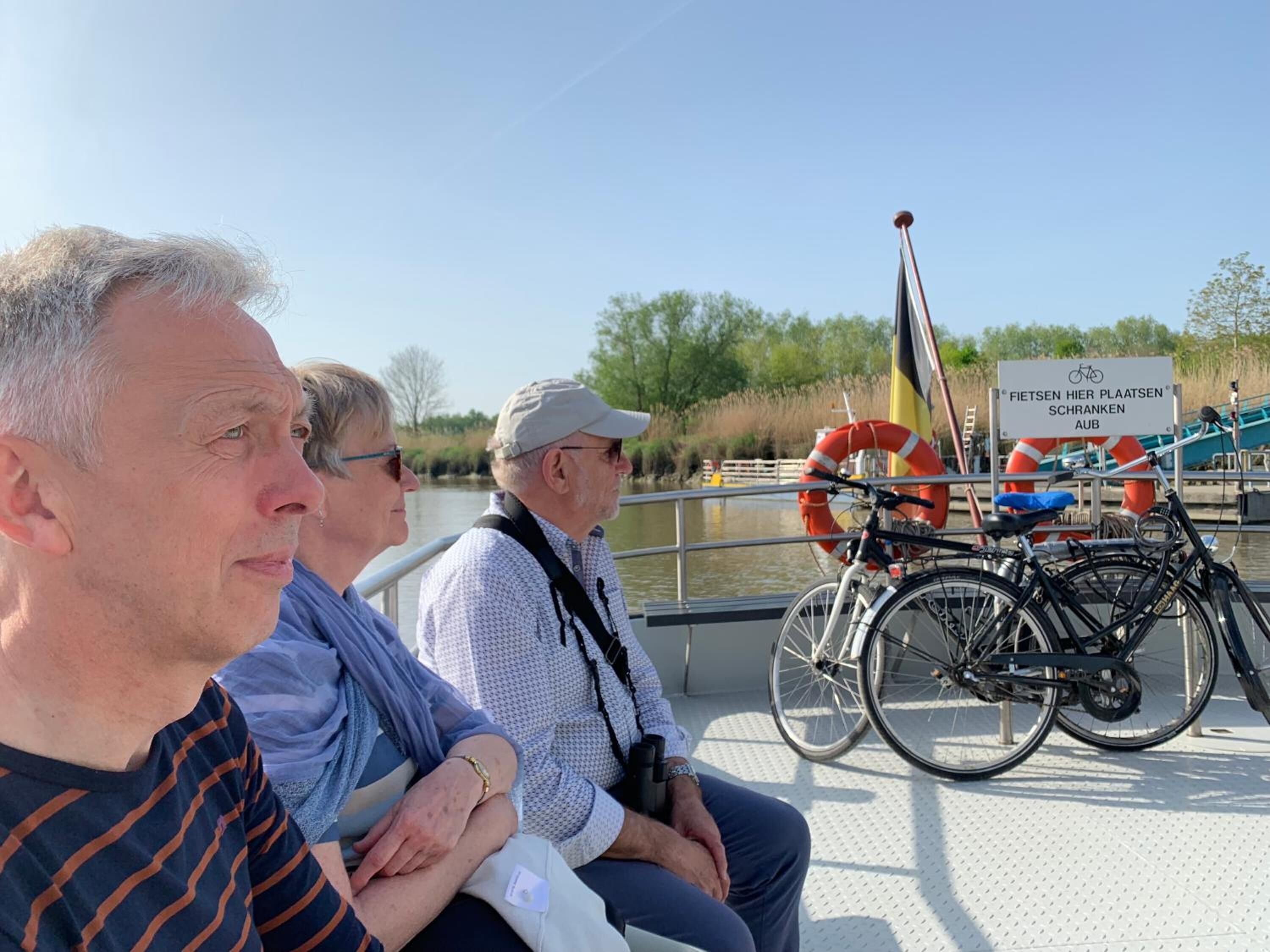 Drie mensen zitten op een boot, met fietsen geparkeerd in de buurt. Een bordje op de achtergrond wijst op een plek die met fietsen te maken heeft.