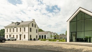 Un grand bâtiment blanc, doté d’une extension moderne en verre, une allée pavée et une voiture garée.