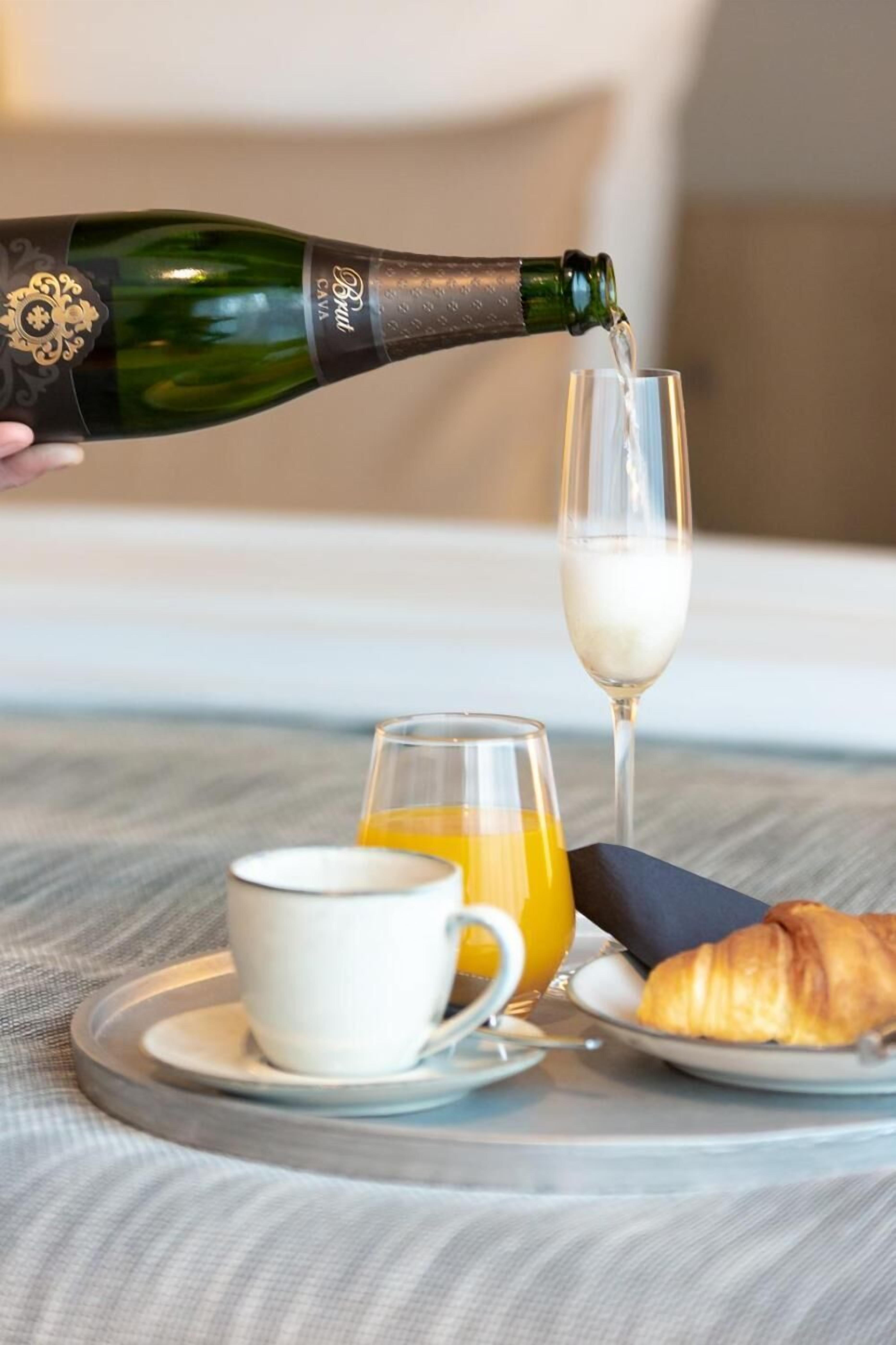 Una bottiglia di champagne che viene versata in un bicchiere, una tazza di caffè, un bicchiere di succo d'arancia e un croissant su un tavolo.