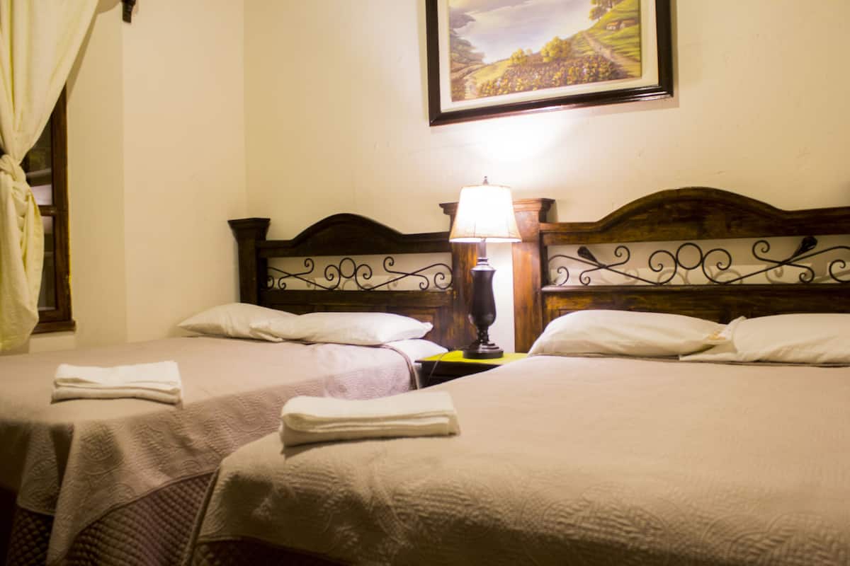 A hotel room with two beds, each with a neatly folded towel, a bedside lamp, and a framed landscape painting on the wall.