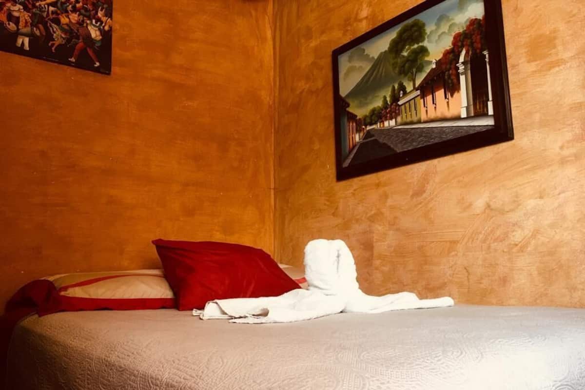 A hotel room with a bed, a red pillow, a white towel, and a framed picture on the wall.
