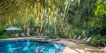 Zona de piscina con sillones de descanso y una mesa bajo una pérgola, rodeada de exuberante vegetación.