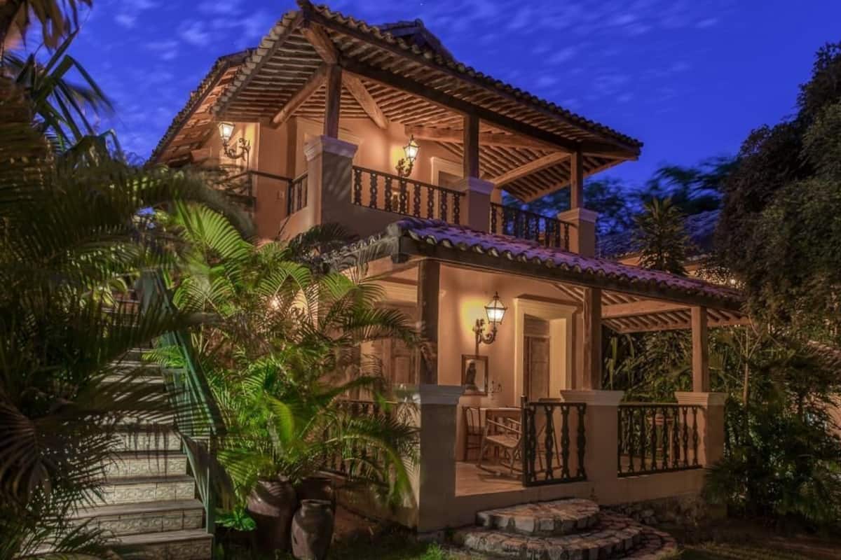A two-story house with a balcony, surrounded by tropical plants.