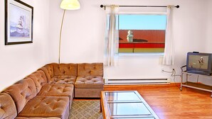 Uma sala de estar com sofá seccional, mesa de centro de vidro, televisão em um móvel, janela grande com cortinas e um quadro na parede.