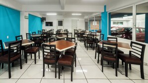 Un restaurante con varias mesas y sillas, suelo de baldosas y vista a coches aparcados a través de amplias ventanas.