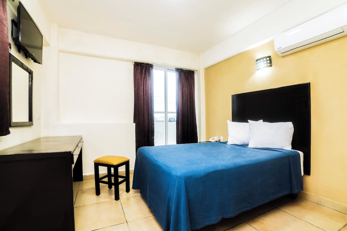 A hotel room with a bed, a desk, a chair, a window with curtains, and an air conditioning unit.