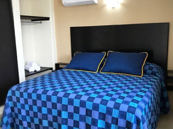 A hotel room with a checkered blue and black bedspread, two matching pillows, a dark headboard, and a white air conditioning unit on the wall.