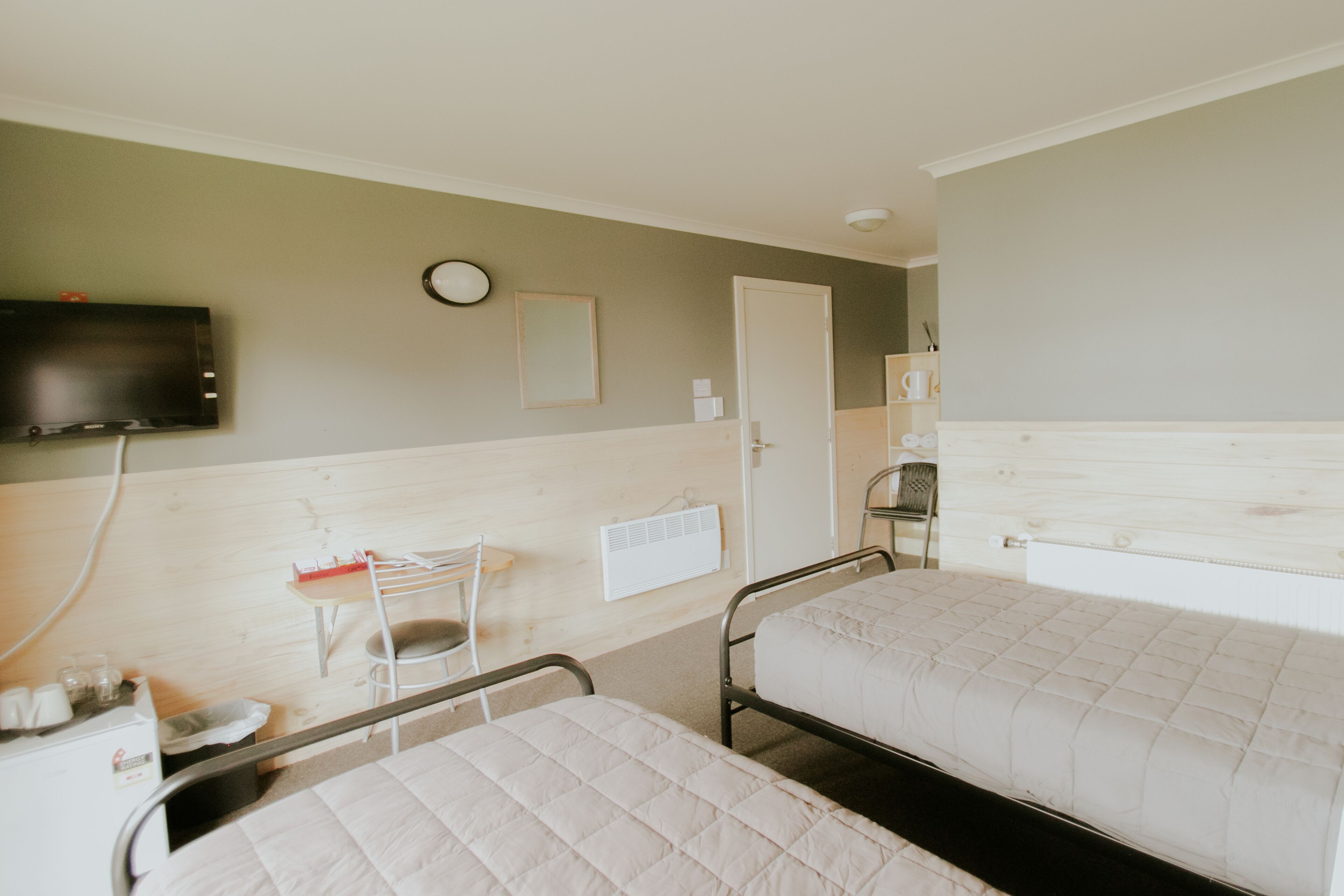 Een kamer met twee bedden, een aan de muur bevestigde televisie, een klein tafeltje met een stoel en een radiator.