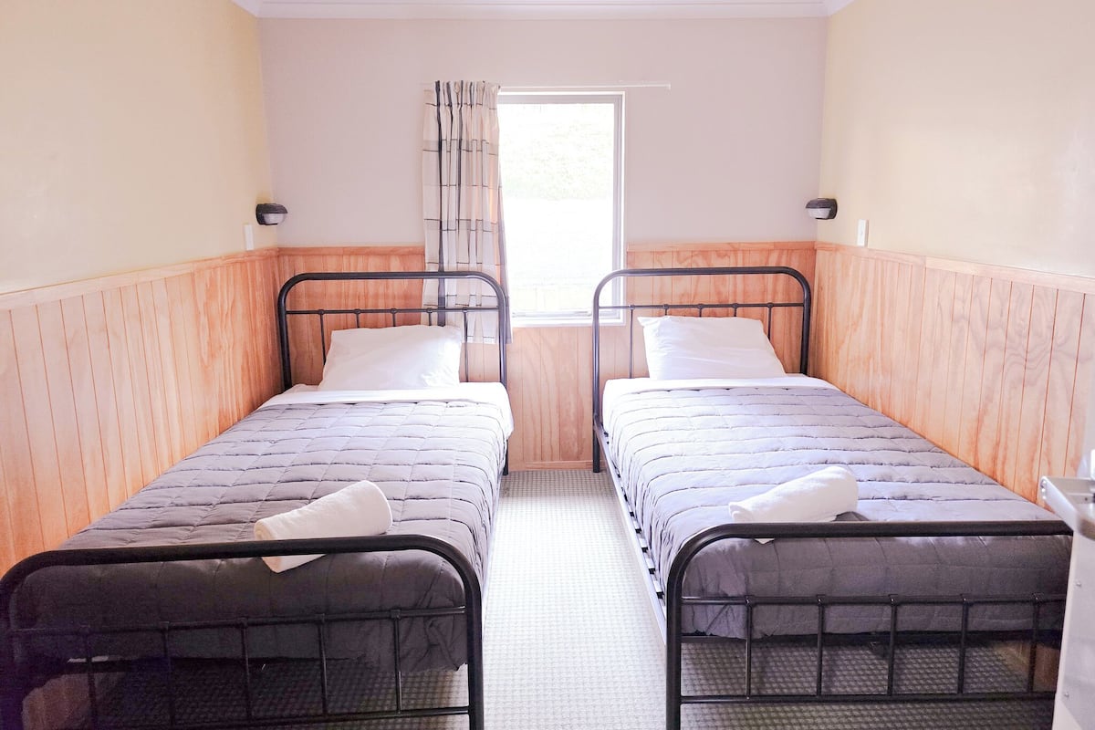 A twin room with two single beds, wooden paneling, a window with curtains, and a small bedside table.