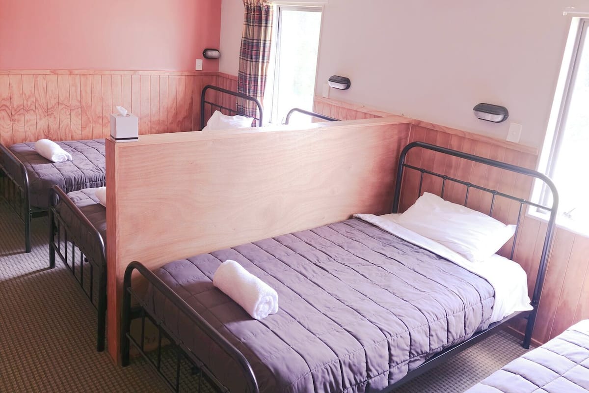 A hotel room with two single beds, a wooden headboard, a window with a curtain, and a wall-mounted light fixture.