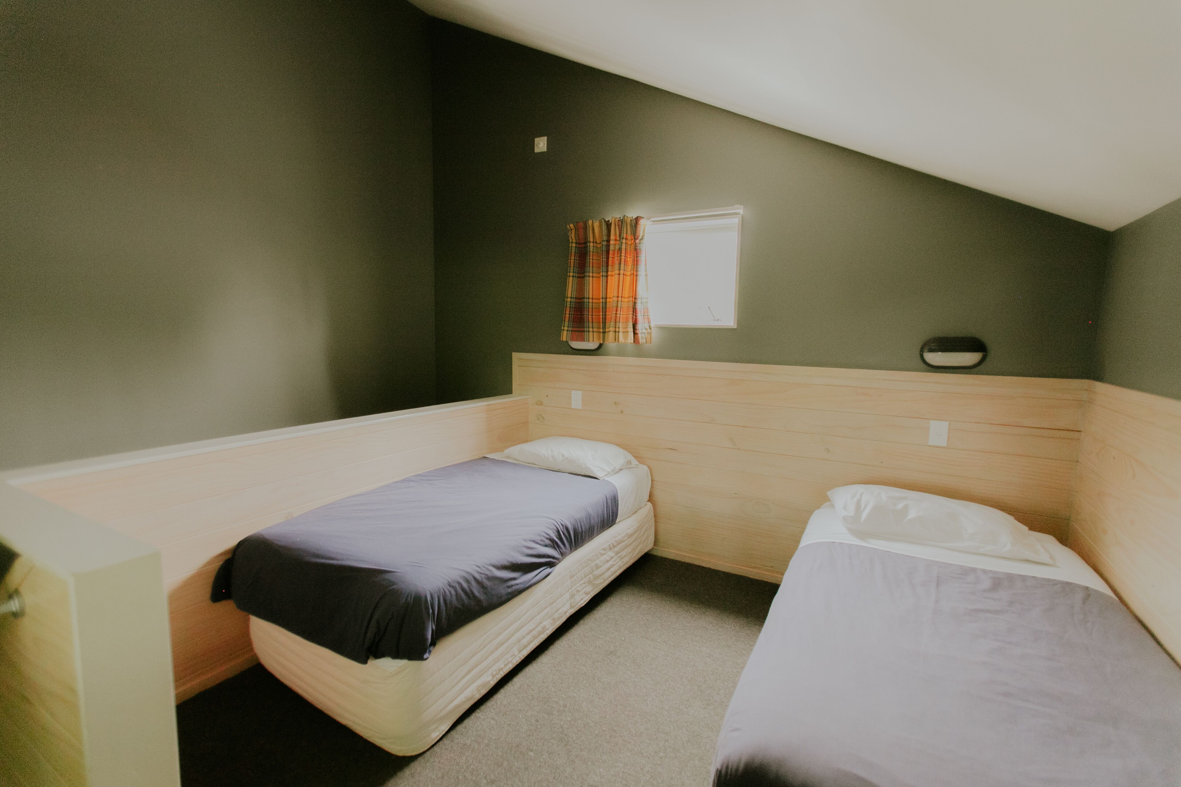 A small, single-room accommodation with two single beds, a window with a curtain, and a wooden headboard.