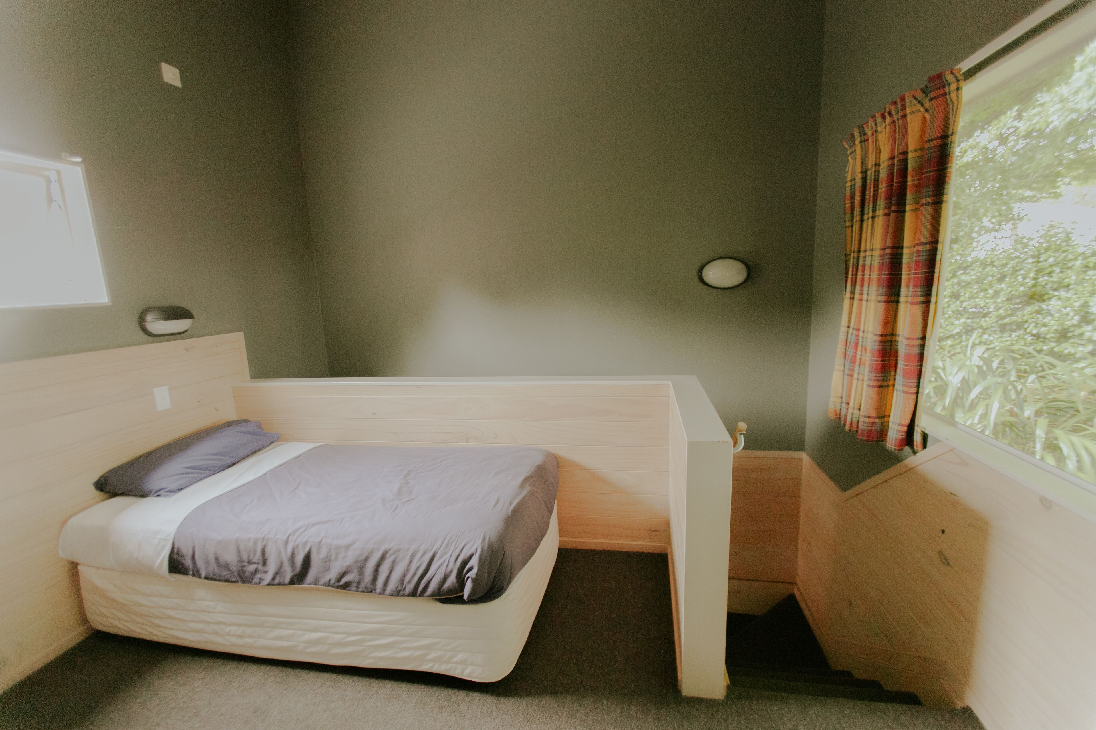 A small, single-bed room with a wooden headboard, a window with curtains, and a small bedside table.