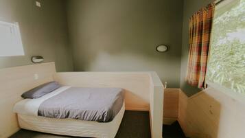 A small, single-bed room with a wooden headboard, a window with curtains, and a small bedside table.
