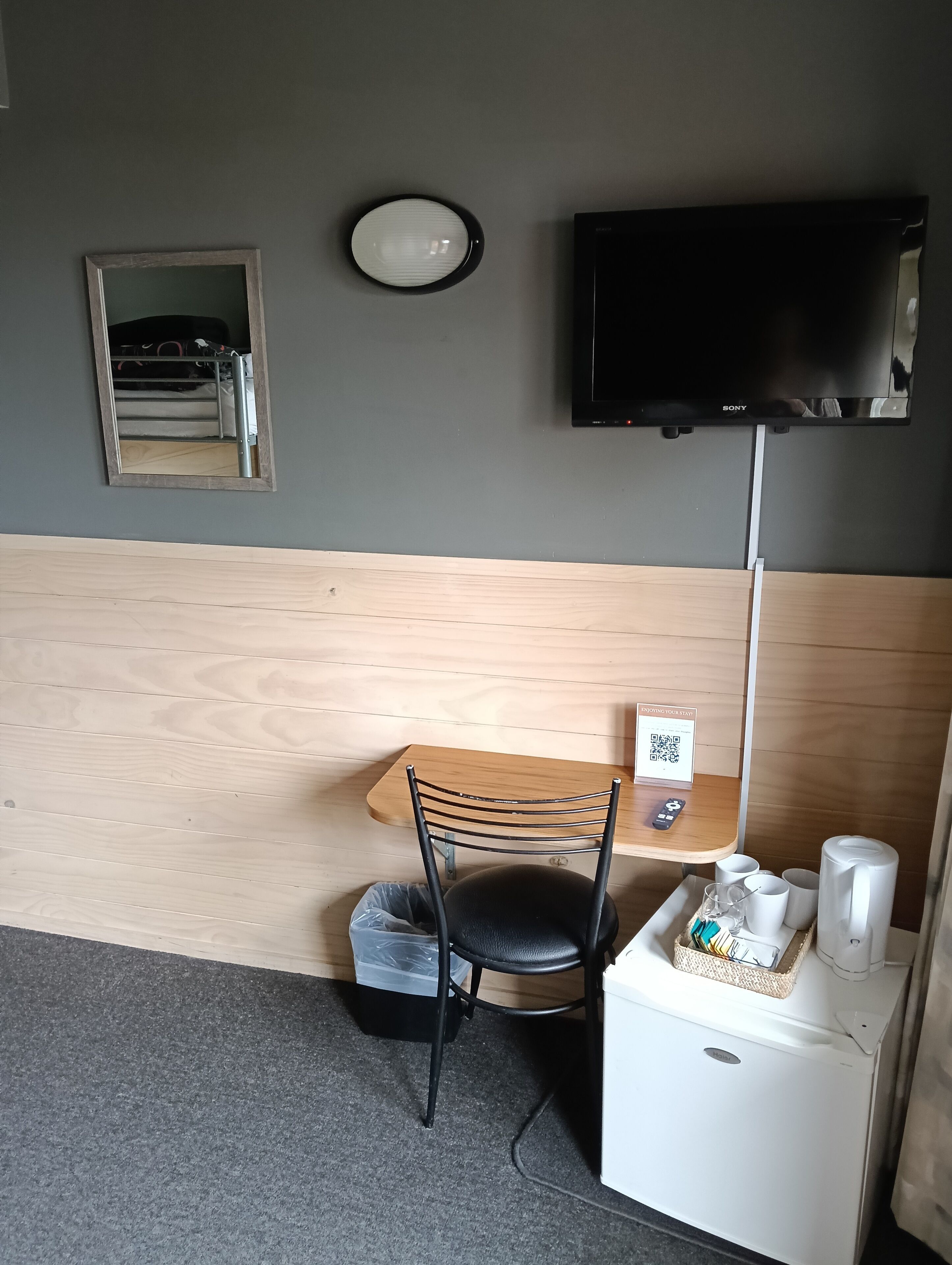 Una habitación pequeña con una mesa de madera, una silla negra, un bote de basura, una máquina de café blanca y un televisor montado en la pared.