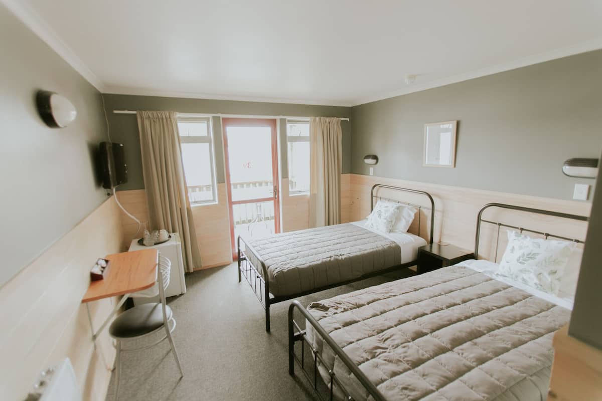 A hotel room with two beds, a desk, a chair, a bedside table, a lamp, and a window with curtains.