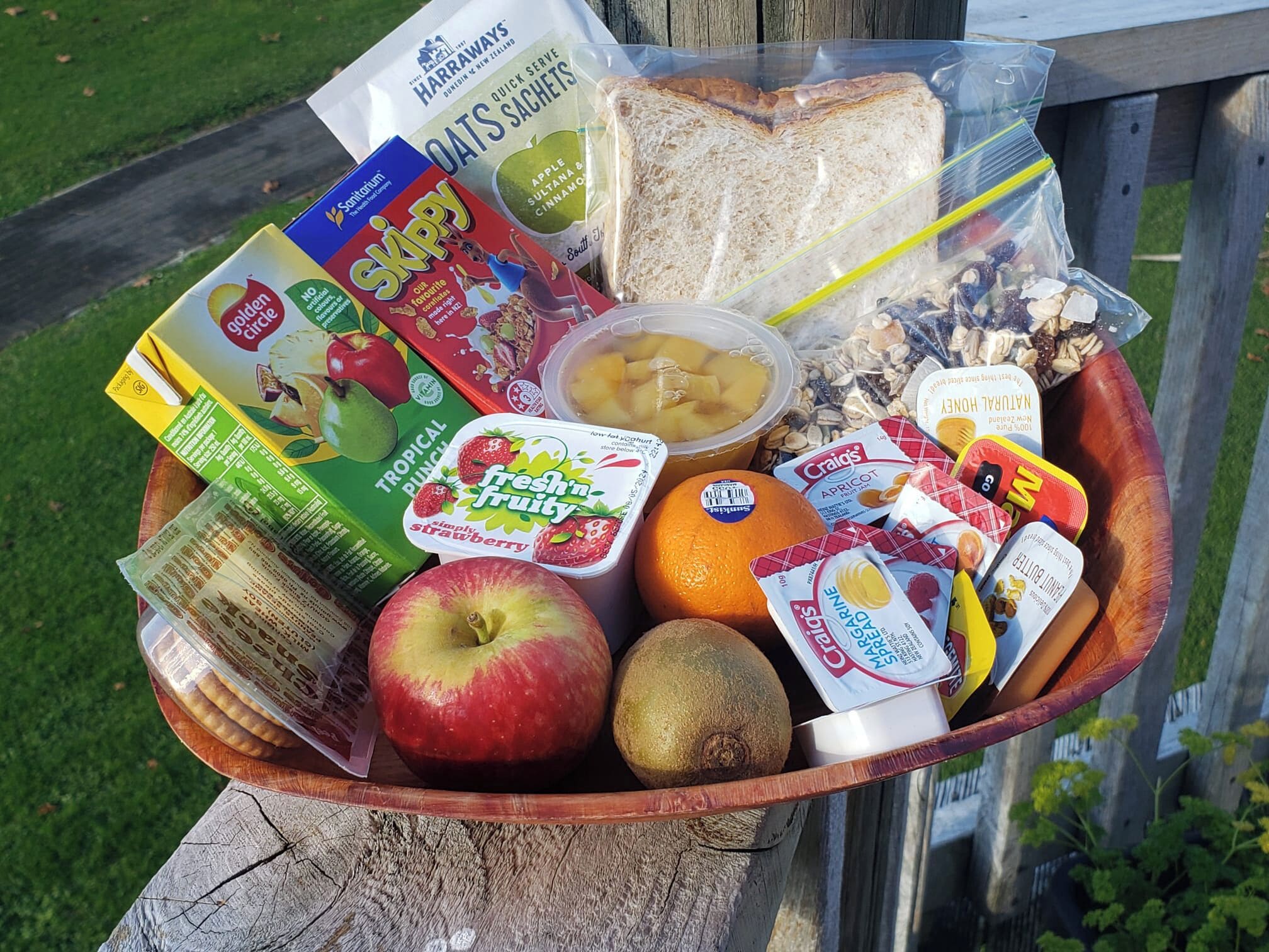 En kurv af træ fyldt med forskellig mad, heriblandt brød, æbler, appelsiner og pakker med snacks.