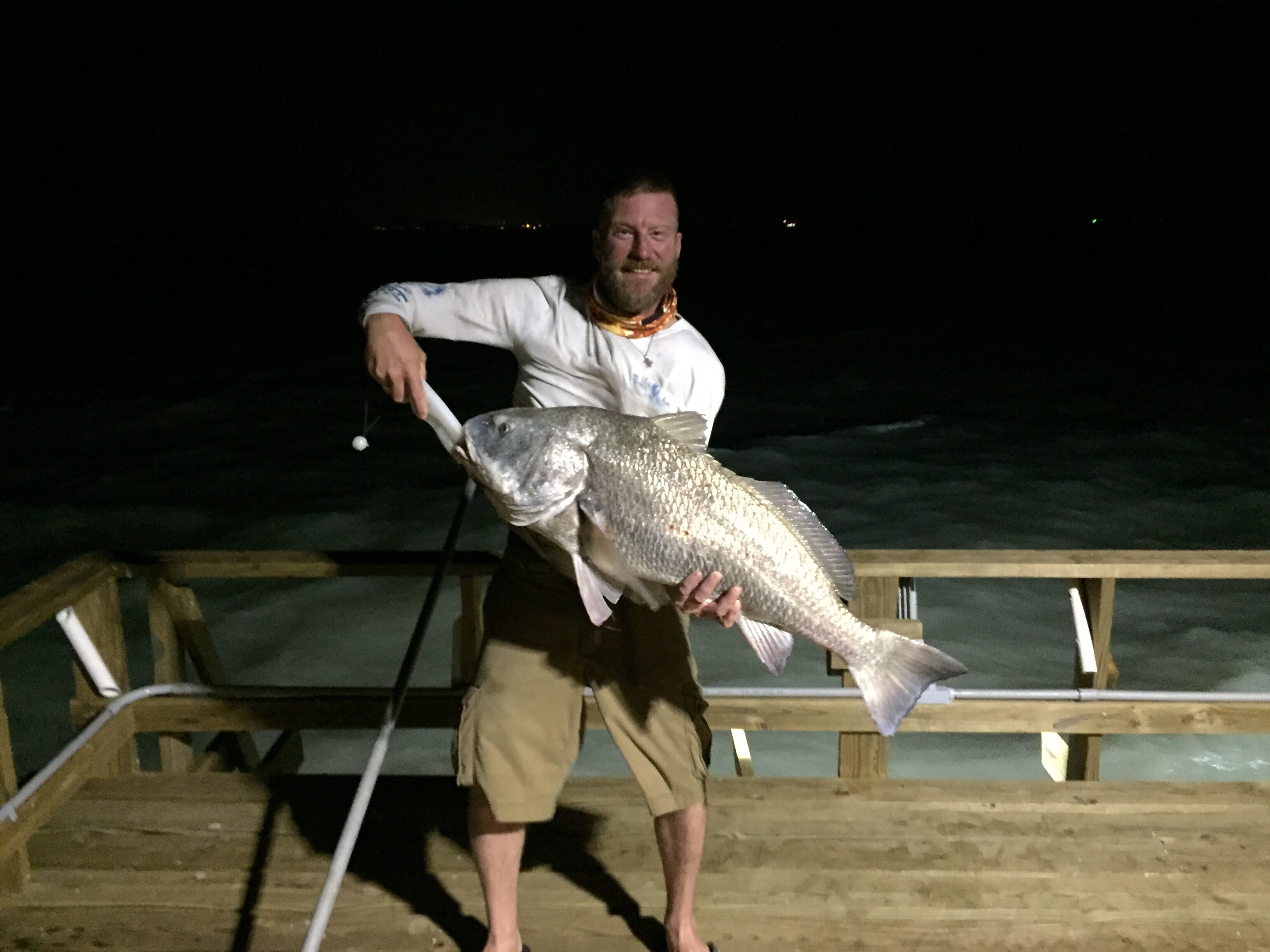 A person holding a large fish on a wooden dock at night.