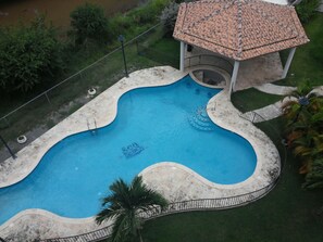 Vue aérienne d’une piscine à la forme incurvée, entourée d’une terrasse en béton et d’un petit pavillon doté d’un toit en tuiles.