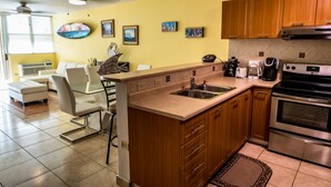 A modern kitchen with wooden cabinets, a stainless steel sink, and a built-in oven. There is a dining area with a glass table and chairs, and artwork on the walls.