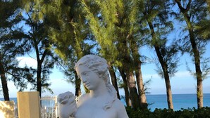 Une statue blanche reprĂ©sentant une femme tenant un panier de fruits, placĂ©e sur un fond composĂ© dâarbres et offrant une vue sur la mer.