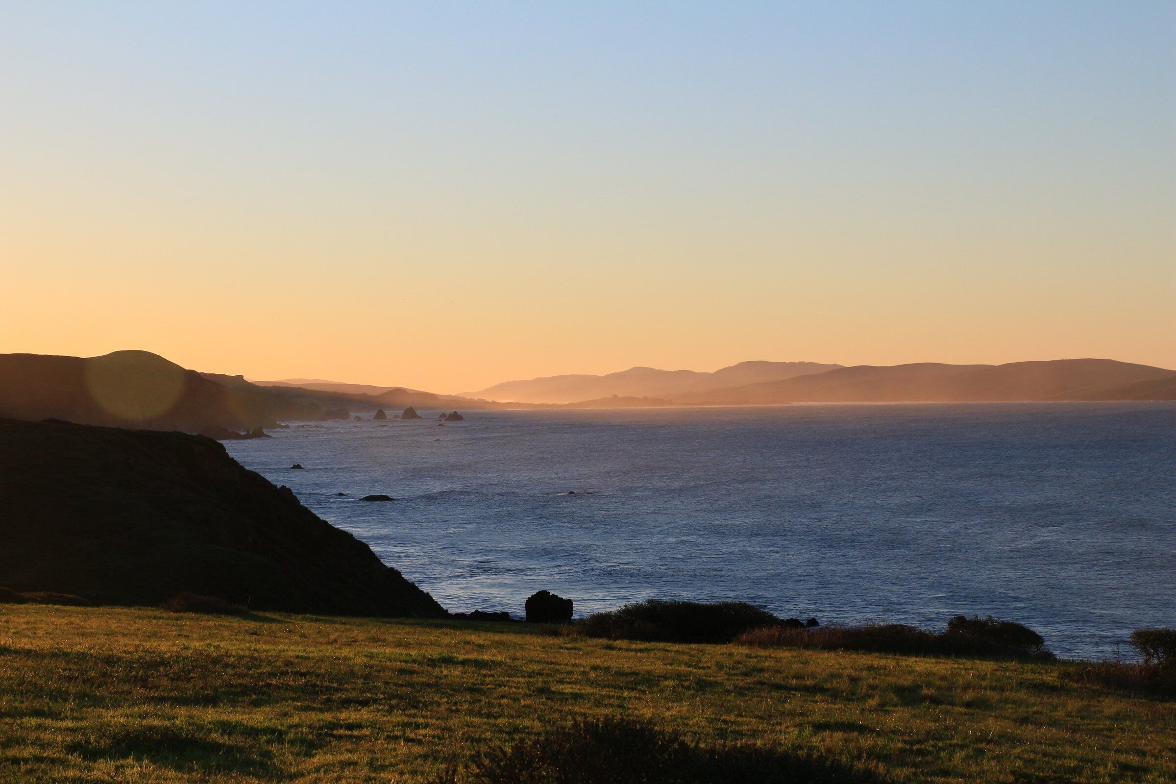 A coastal landscape with a grassy foreground and a view of the sea and distant mountains.