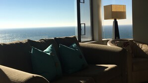 Ein Wohnzimmer mit einem großen Fenster mit Meerblick, einem Sofa mit türkisfarbenen Kissen und einer Stehlampe.