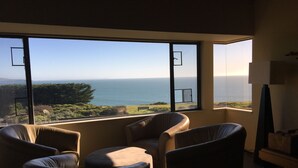 Ein Zimmer mit einem großen Fenster, das Blick auf den Ozean und die Hügel bietet. Drei Ledersessel und eine Lampe stehen auf einem Tisch.