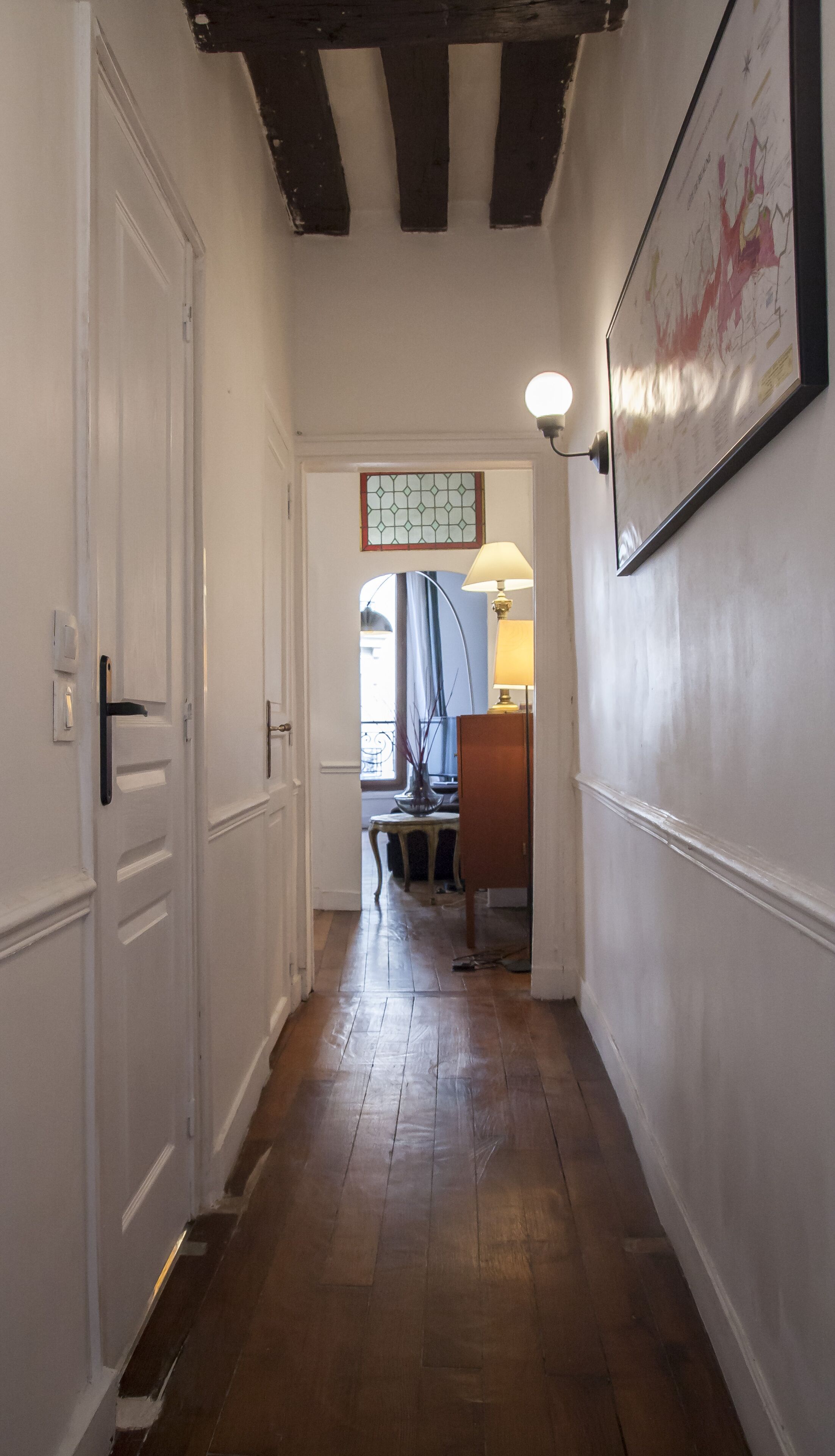 Ein Flur mit Holzboden, weißen Wänden und einer Decke mit freiliegenden Balken. Links eine Tür, rechts eine Lampe und ein gerahmtes Bild an der Wand.