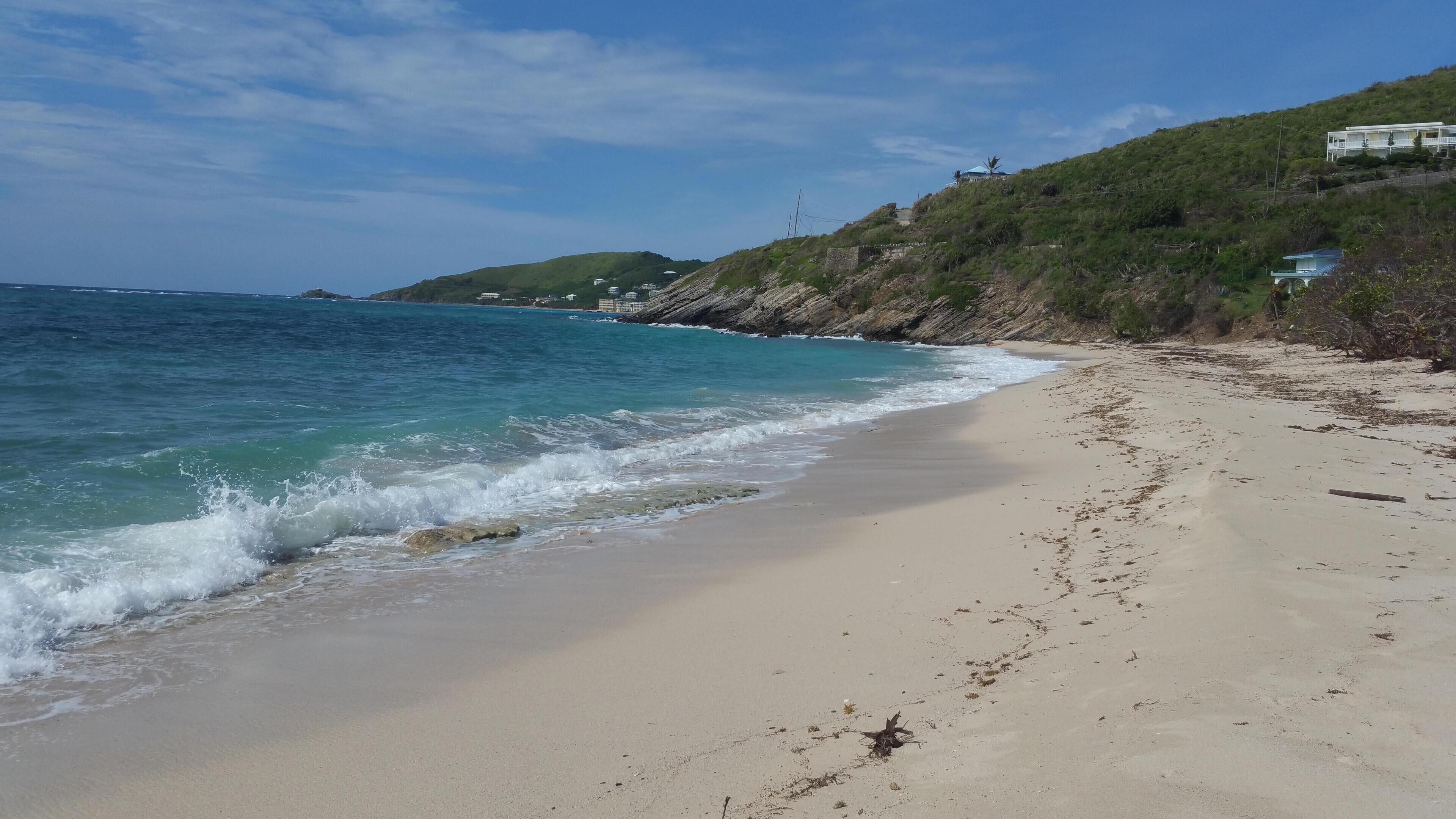 Ein Strand mit klarem, blauem Wasser, sandigem Ufer und einem Gebäude auf einem Hügel im Hintergrund.