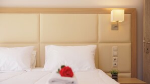 Un lit bien fait, avec du linge de lit blanc, une rose rouge et une serviette pliée posée sur le lit. Une petite plante en pot se trouve sur la table de chevet, à côté du lit.