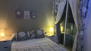 Ein ordentlich bezogenes Bett mit gemusterten Bettwäsche, ein Nachttisch mit Lampe und ein Fenster mit Vorhängen.