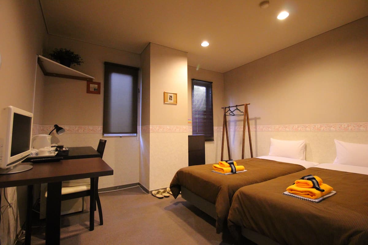A hotel room with two beds, a desk with a computer, a chair, and a small shelf with a plant.