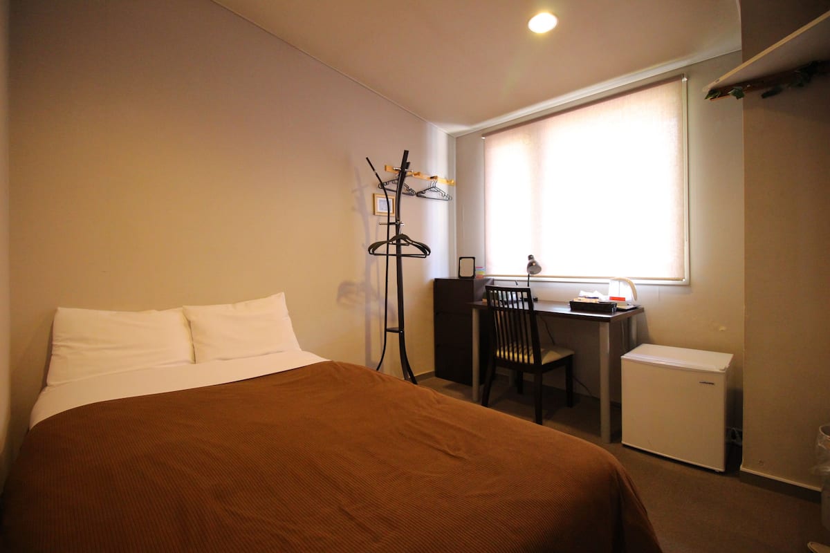 A hotel room with a bed, a desk, a chair, a small refrigerator, and a coat rack.