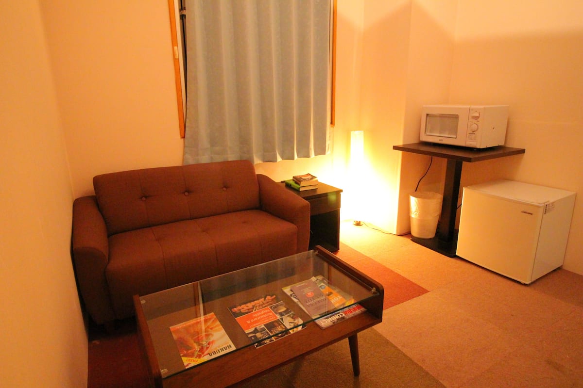 A living room with a brown sofa, a glass-top coffee table with magazines, a white microwave, and a small refrigerator.