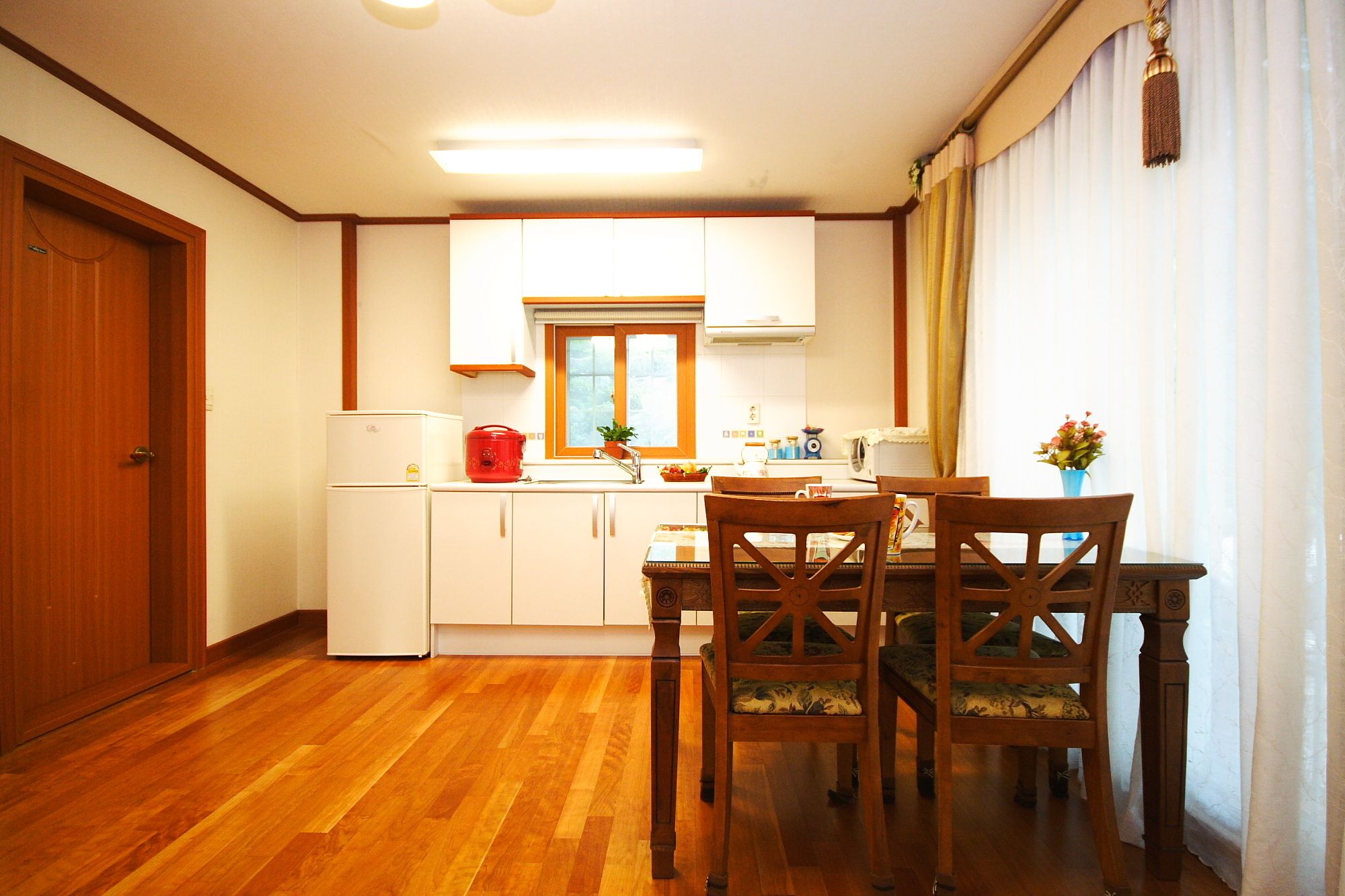 A compact kitchen with wooden cabinets, a dining table with chairs, and a window with curtains.