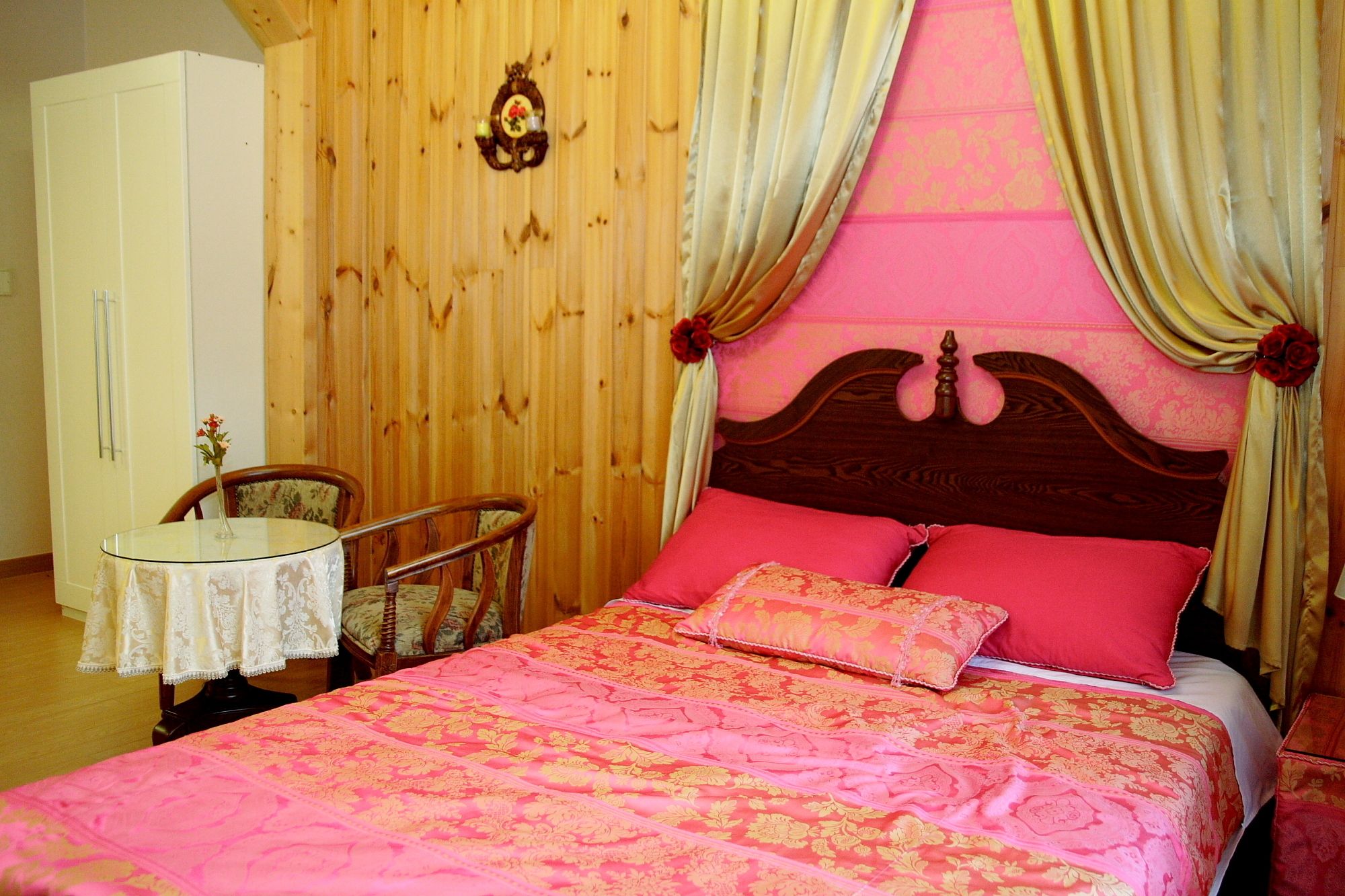 A bedroom with a wooden headboard, a bed with pink and gold bedding, a small round table with a lace tablecloth, and a wooden wardrobe.